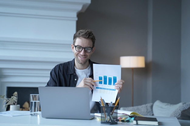 Young smiling german businessman pointing at document with graphics during internet meeting