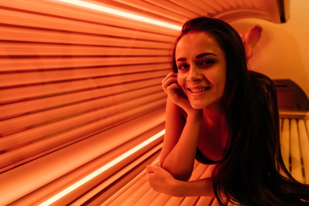 A young smiling girl lies in a tanning bed under the ultraviolet rays, sunbathing, wants a tanned skin