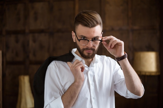 Young smiling handsome bearded businessman in a white shirt and glasses stands at the center of the office