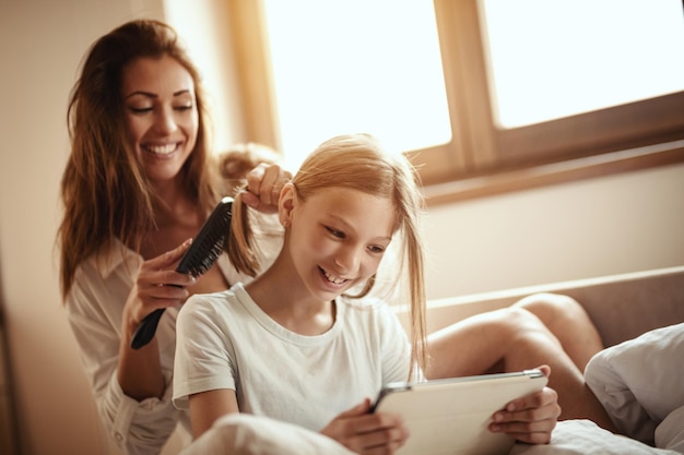 Young smiling happy mother is combing her little daughter's hair on bed while the girl is looking at the digital tablet.