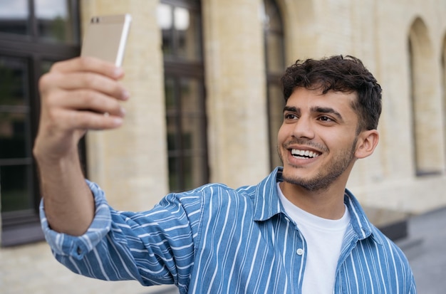 Young smiling Indian man holding mobile phone taking selfie on the street