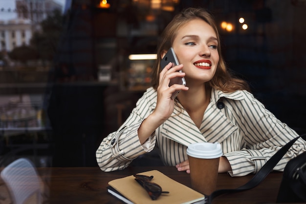 Young smiling lady in striped trench coat talking on cellphone happily 