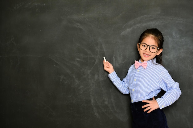 young smiling little teacher standing in blackboard background and looking at camera ready to using chalk teaching during school class.