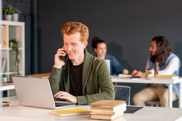 Young smiling male office manager with smartphone looking at laptop display while talking to clients by phone and answering their questions