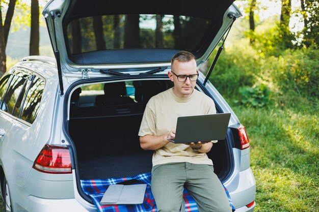 Young smiling man in glasses freelancer works on a laptop while sitting in the trunk of a car Remote work in nature