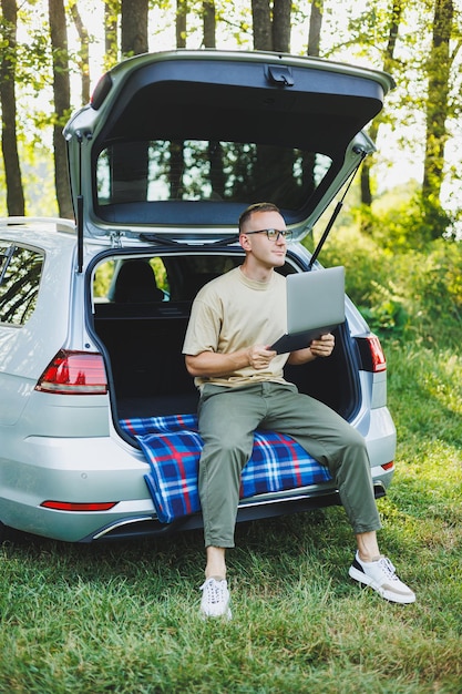 Young smiling man in glasses freelancer works on a laptop while sitting in the trunk of a car Remote work in nature