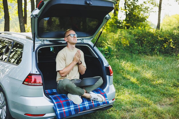 Young smiling man in glasses freelancer works on a laptop while sitting in the trunk of a car Remote work in nature