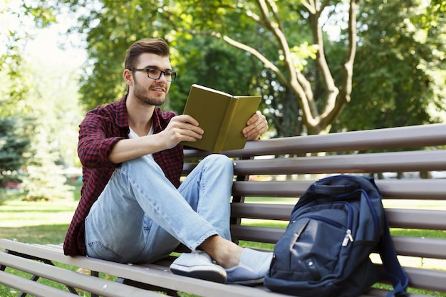 Young smiling man in glasses reading book in park, studying and preparing for exams at university or college. Technology, communication, education and remote working concept, copy space