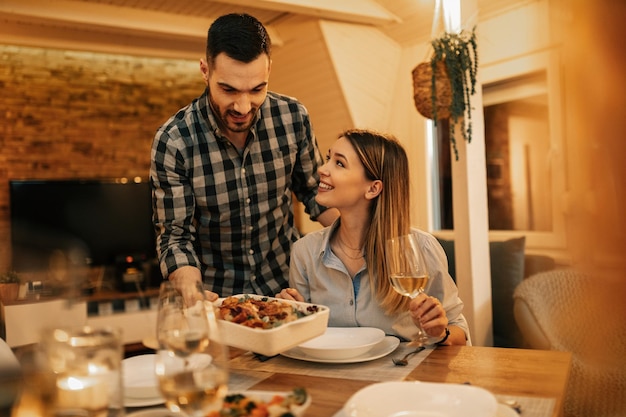 Young smiling man serving dinner to his girlfriend in dining room