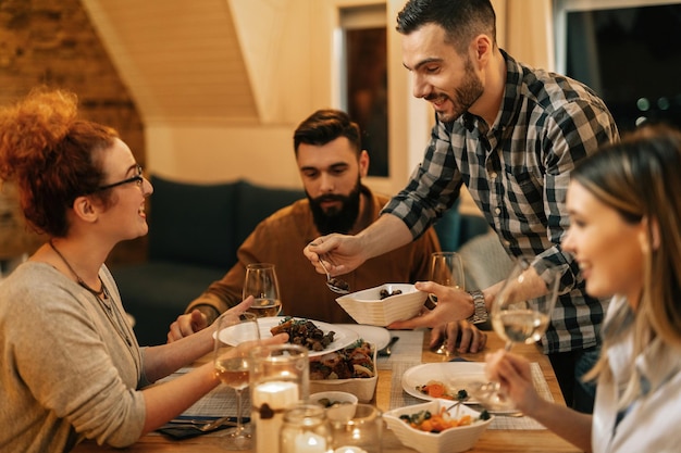 Young smiling man serving food while having dinner with his friends at dining table at home