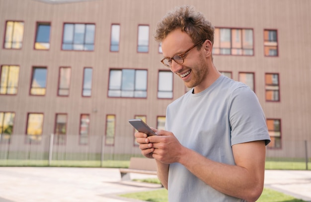 Young smiling man using mobile phone shopping online ordering something standing on the street