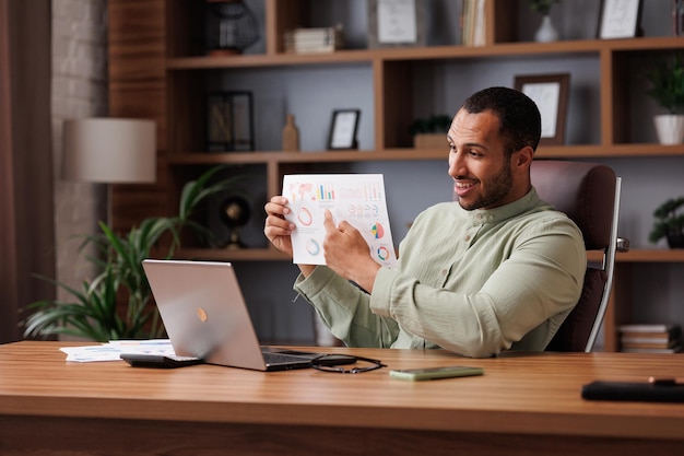 Young smiling mixed race businessman have video call sitting in office showing financial report with graphs and charts to colleagues partners and investors Online video conference