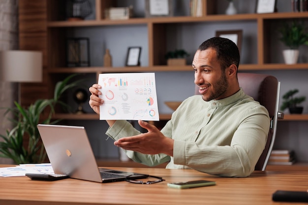 Young smiling mixed race businessman have video call sitting in office showing financial report with graphs and charts to colleagues partners and investors Online video conference