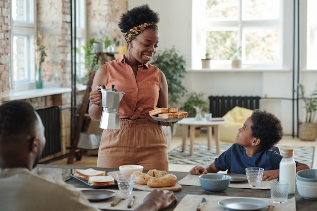 Young smiling mother in casualwear passing homemade waffles to her cute little son going to have breakfast with his parents in the kitchen