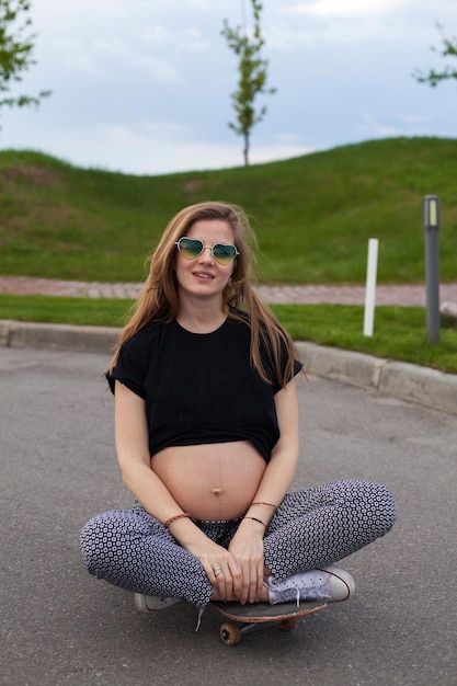 Young smiling pregnant woman in black Tshirt and heartshaped sunglasses sitting on the skateboard on the road on the background of green lawn