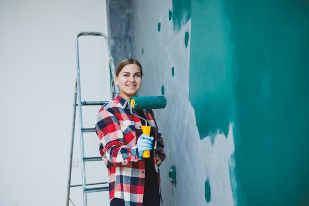 Young smiling pretty woman in shirt holding roller in right hand and painting wall Repair in the room