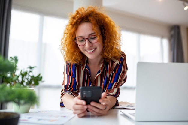 Young smiling red hair business woman using smartphone near lptop in office