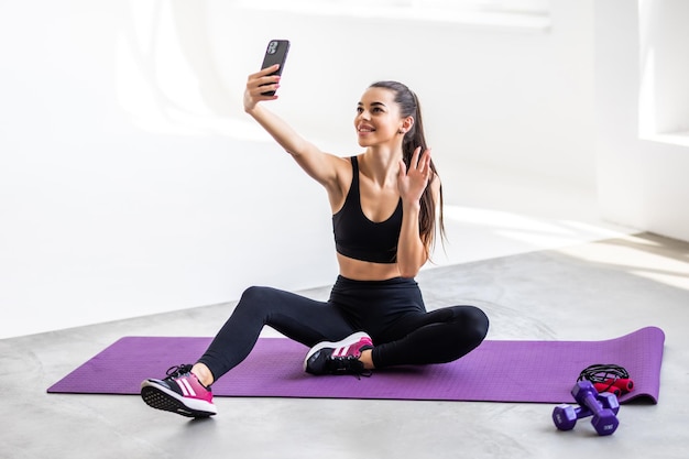 Young smiling strong sporty athletic fitness trainer instructor woman doing exercises do selfie shot on mobile cell phone sit on mat floor at home gym