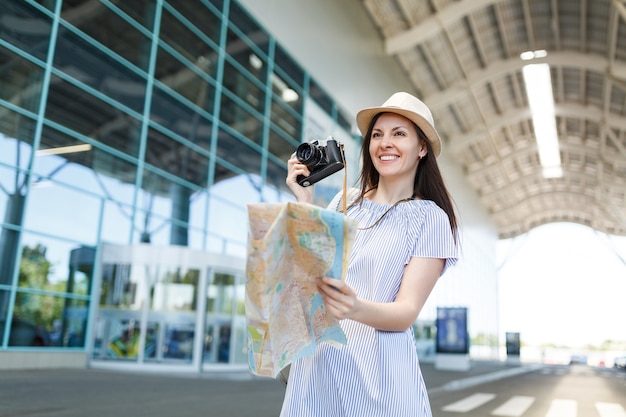 Young smiling traveler tourist woman in hat holding retro vintage photo camera, paper map at international airport