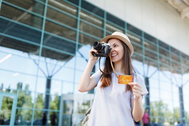 Young smiling traveler tourist woman in hat, take pictures on retro vintage photo camera, hold credit card at international airport