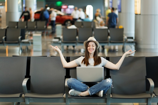 Young smiling traveler tourist woman with laptop sitting with crossed legs, meditate, spread hands, waiting in lobby hall at airport