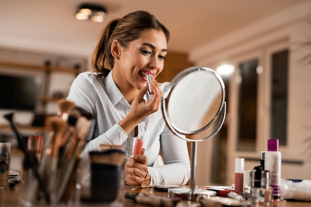 Young smiling woman applying lipstick while looking herself in a makeup mirror