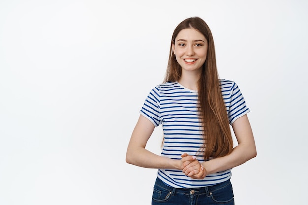 Young smiling woman asissting, holding hands together and looking friendly at camera, posing against white background.