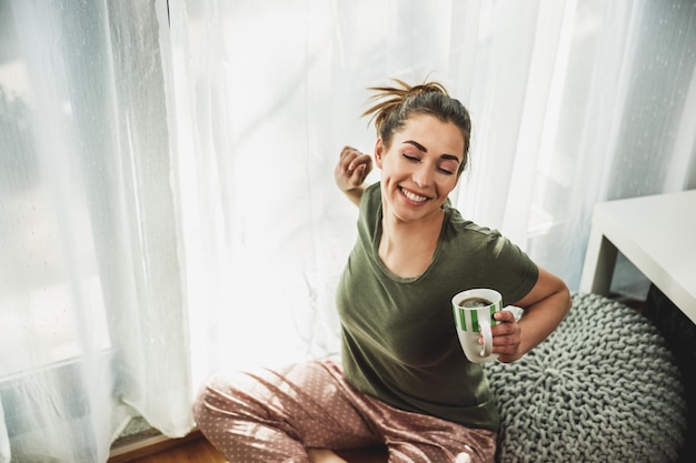 Young smiling woman enjoying coffee in the cozy morning sunshine near the window at home or hotel room.