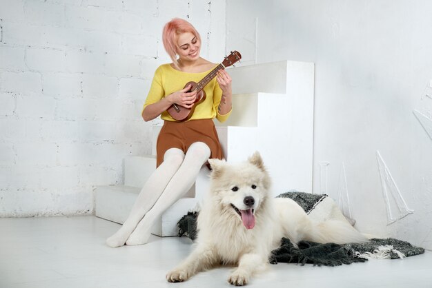 Young smiling woman girl in skirt and blouse, playing the ukulele, sitting on the steps with a smile on her face, while her white dog Samoyed lying on the rug at her feet.