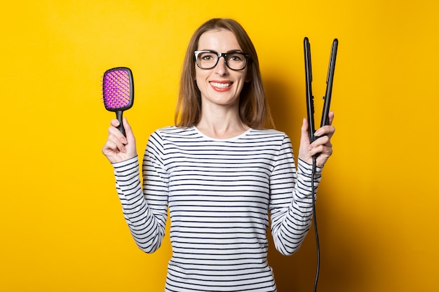 Photo young smiling woman holds a hairbrush and curlers for hair on a yellow background