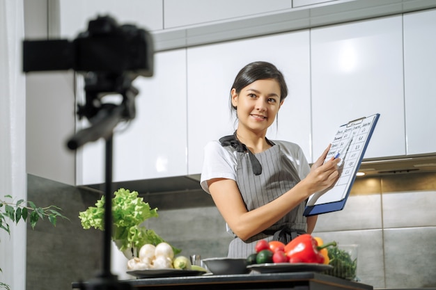 Young smiling woman nutritionist showing a comparative table of calorie foods and recording video on a camera in the kitchen.