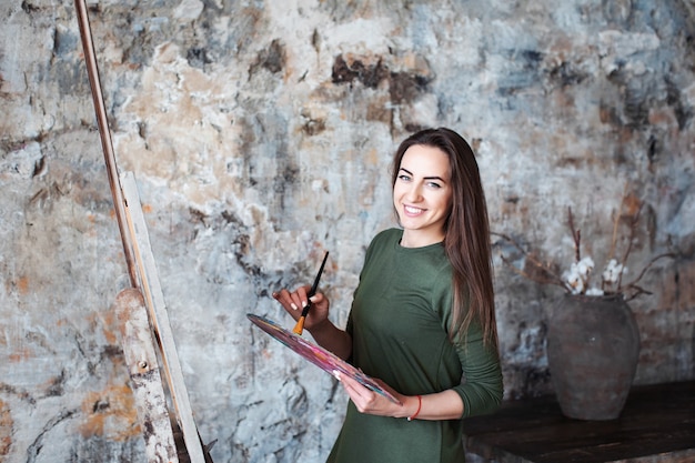 Photo young smiling woman painter in a green dress with a brush and a palette in an art studio. hobby.