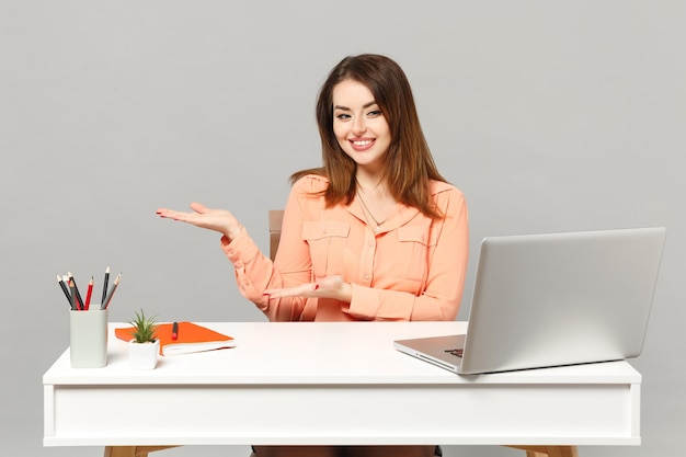 Young smiling woman in pastel casual clothes pointing hands aside, sit and work at desk with pc laptop isolated on gray background. Achievement business career lifestyle concept. Mock up copy space.