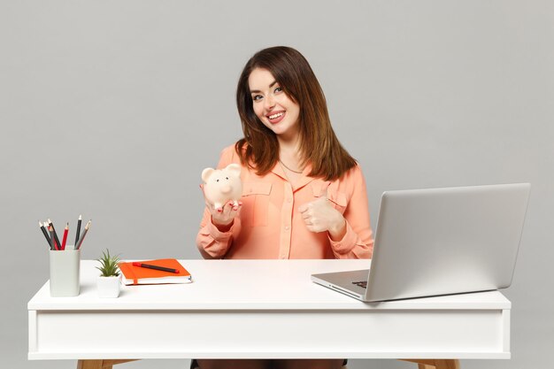 Young smiling woman in pastel clothes showing thumb up hold piggy money bank work at desk with pc laptop isolated on gray background. Achievement business career lifestyle concept. Mock up copy space.