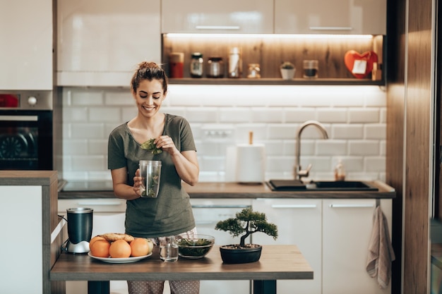 Young smiling woman putting a variety of  vegetables into a blender and making a healthy smoothie at home.