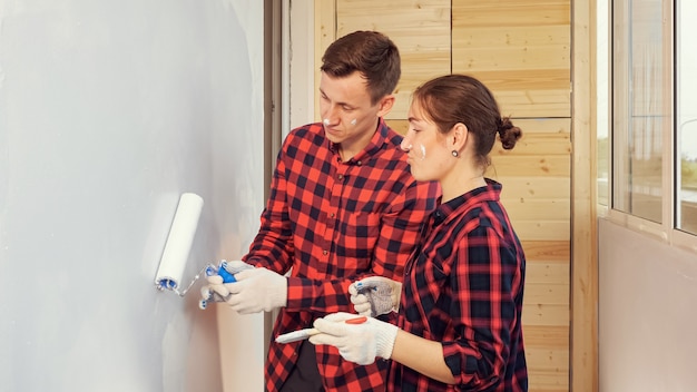 Young smiling woman in red checkered shirt helps cheerful husband paint wall in grey color at repair on light glazed loggia at home