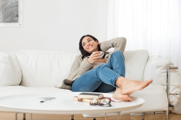 Young smiling woman sitting on sofa and looking up while drinking hot tea