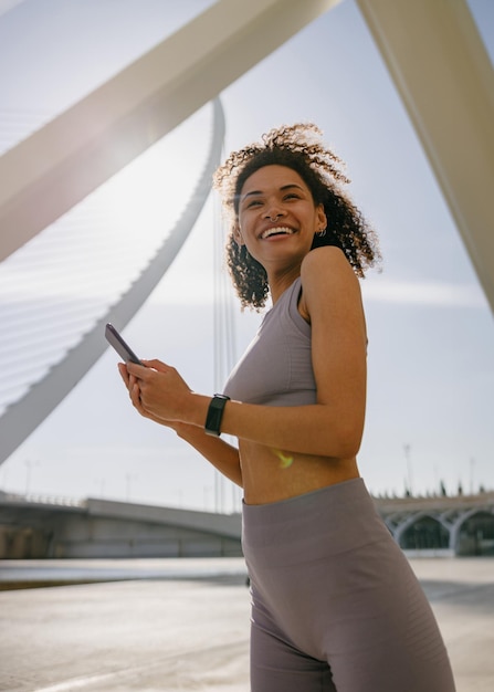 Young smiling woman in sportswear is resting after a workout outside while standing with smartphone