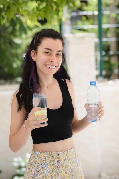 Young smiling woman staying outdoor holding a glass of lemon water in one hand and a bottle of water in another looking to the camera