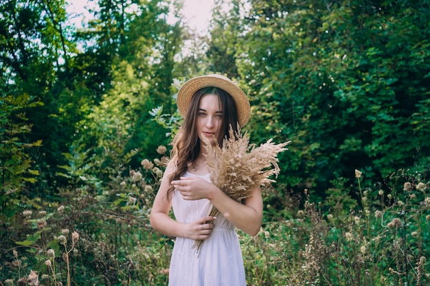 Young smiling woman in a straw hat on a background of green summer forest. Beautiful girl with a bouquet of dried flowers on a walk. Girl in a summer dress.