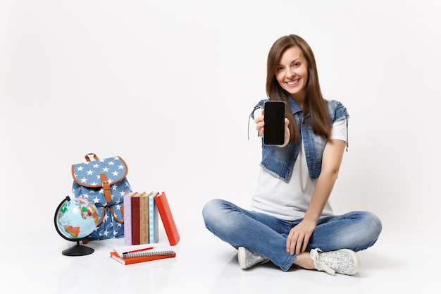 Young smiling woman student holding mobile phone with blank black empty screen, sitting near globe, backpack, school books isolated