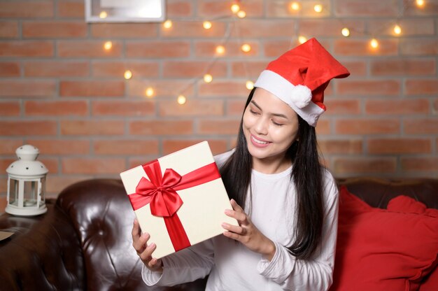 A young smiling woman wearing red Santa Claus hat showing a gift box on Christmas day