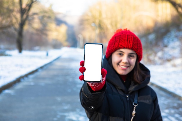 Young smiling woman in winter outfit holding phone with white blank empty screen