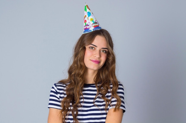 Young smiling woman with long hair wearing in stripped T-shirt and celebration cap on grey background in studio