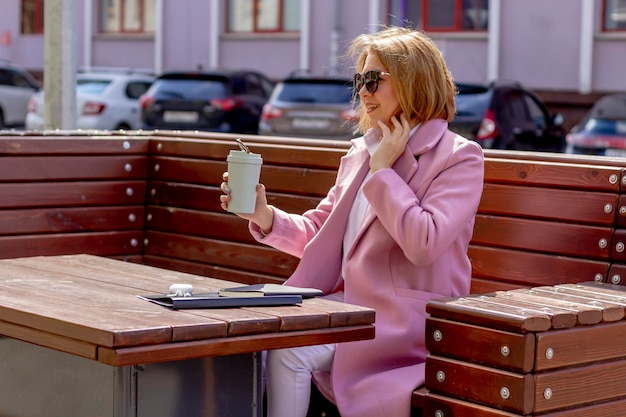 A young smiling womangirl drinks morning coffee resting on a bench against the background of a noisy city