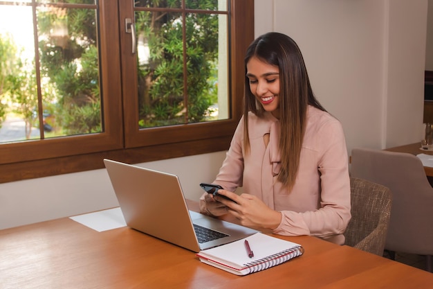 Young smiling working woman using her cell phone while correcting calculations on laptop in a restaurant.