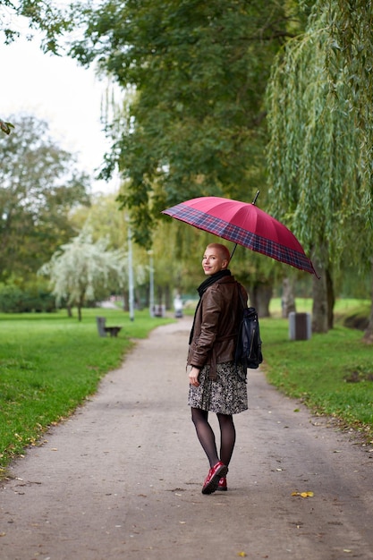 Young smilling short haired blonde woman walks in the autumn park with red umbrella View from back