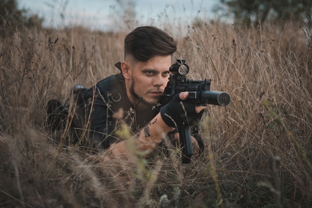 Young soldier in black uniform lying down and aiming an assault rifle