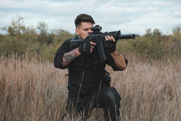 Young soldier in black uniform sitting and aiming with an assault rifle