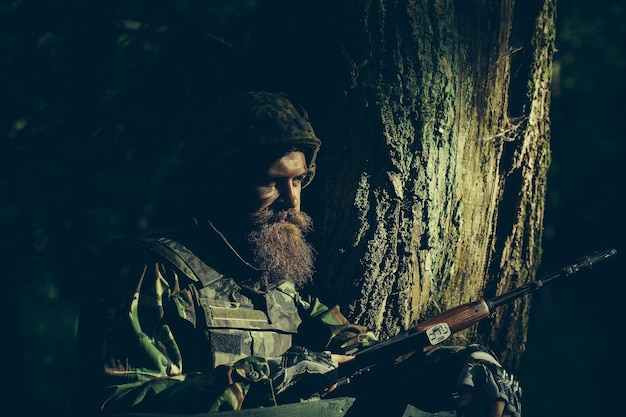 Young soldier with beard on dirty tired face in military uniform and helmet with rifle on tree and g